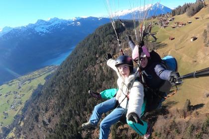 Student paragliding over a steep green hill with snowy mountains in the background.