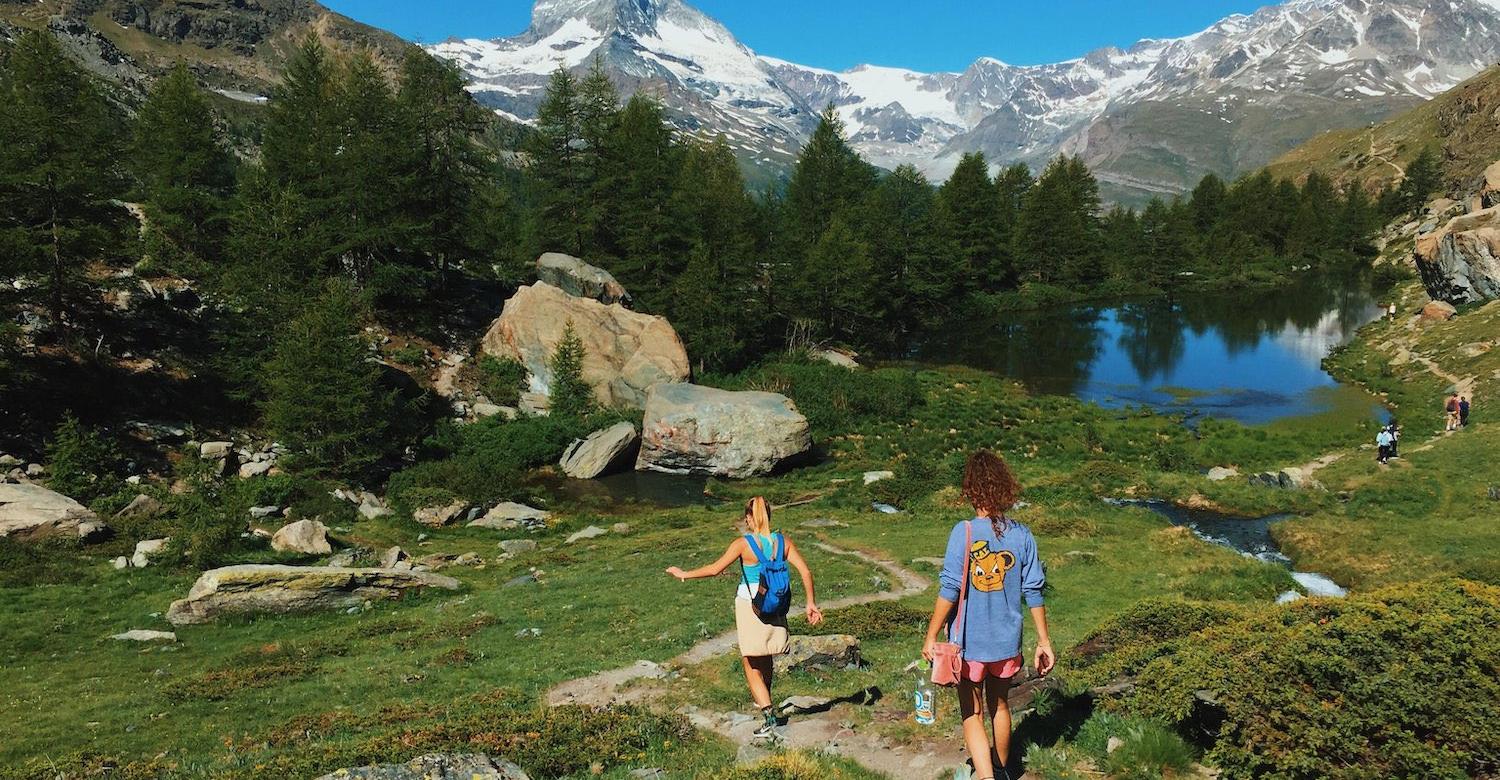 Two hikers on a trail towards a lake with mountains in the background